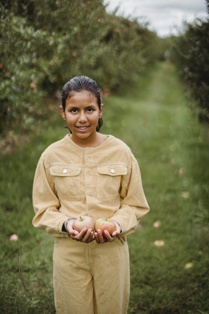 Positive Hispanic girl holding apples in hands while harvesting fruits in farm in countryside
